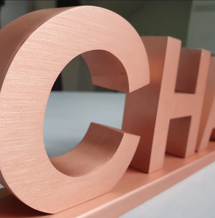 Close das letras caixa CHA em acabamento rose gold acetinado, mostrando volume e textura do metal